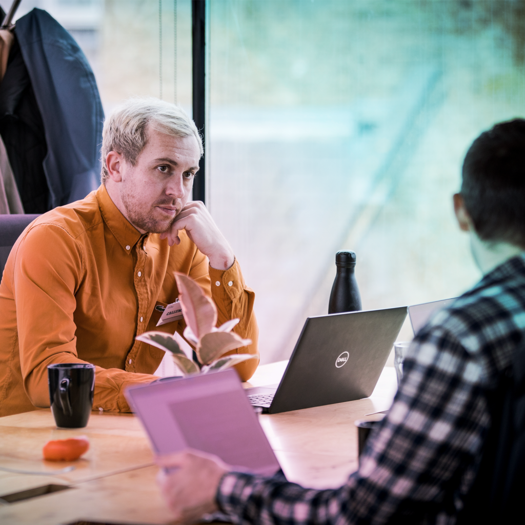 man in an orange-brown shirt sitting at a table, looking intently at another person, listening