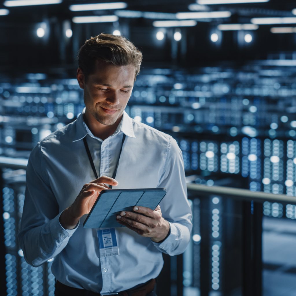 a man looks at a tablet, in the background is a large room of servers.