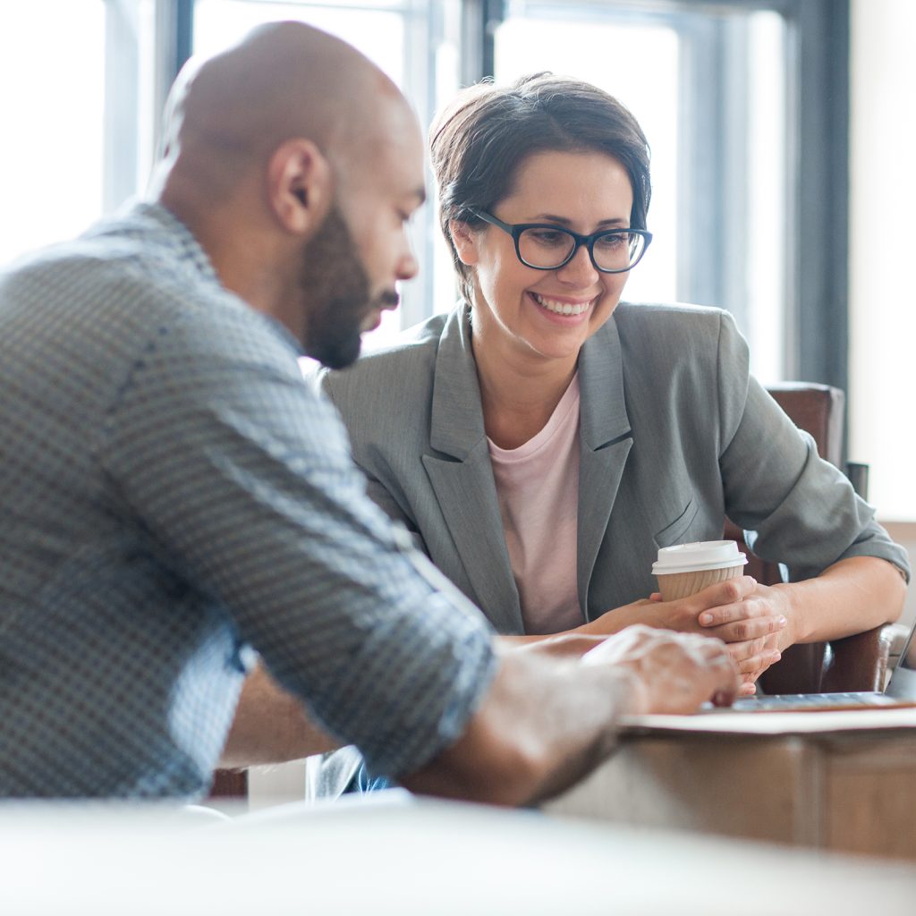 two people dicussing something on a laptop, the woman is holding a coffee